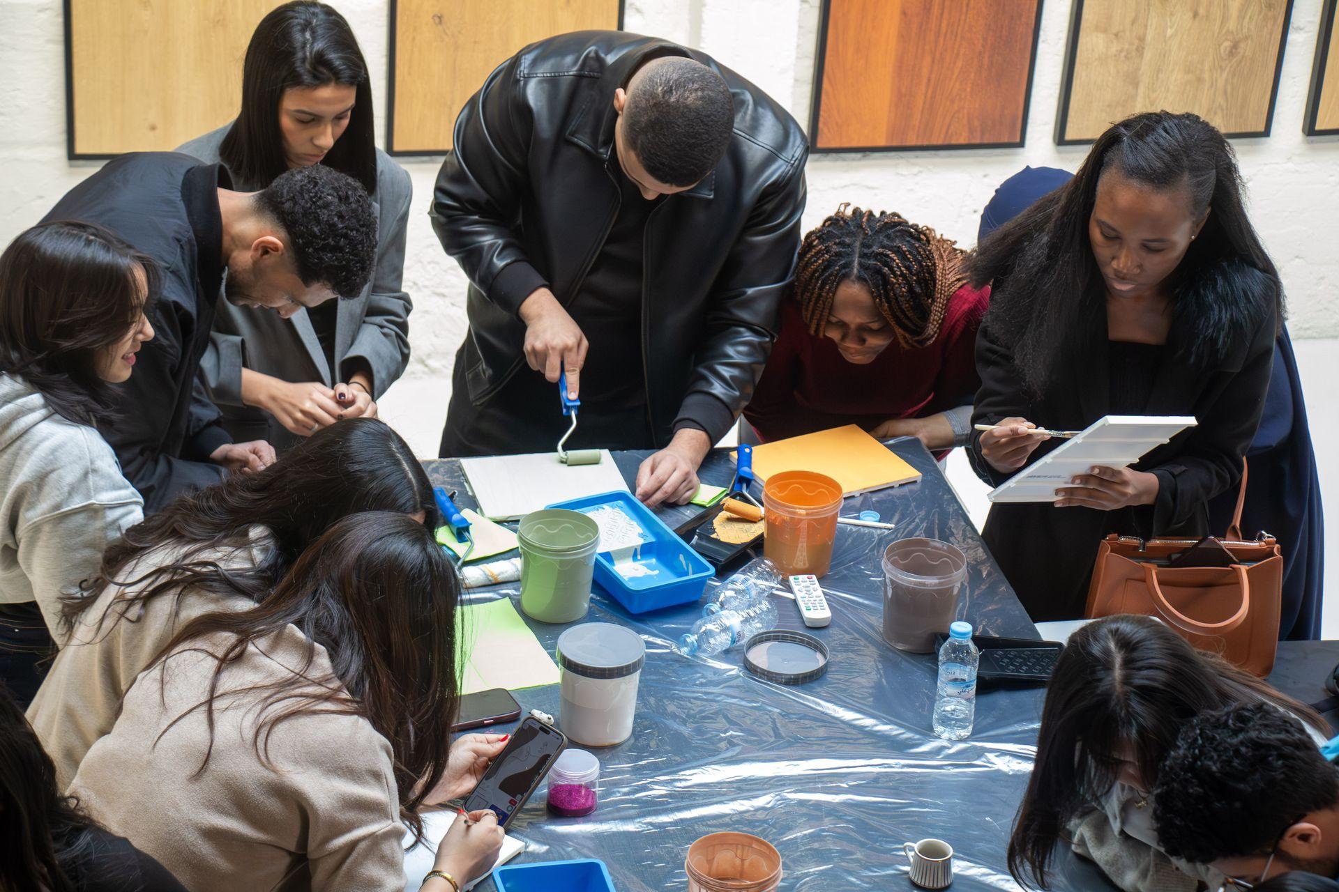Gros plan sur les mains de participants travaillant sur un projet créatif. On voit des pots de peinture (marron et orange), des pinceaux et des paillettes, disposés sur une table recouverte d'une bâche plastique de protection.
